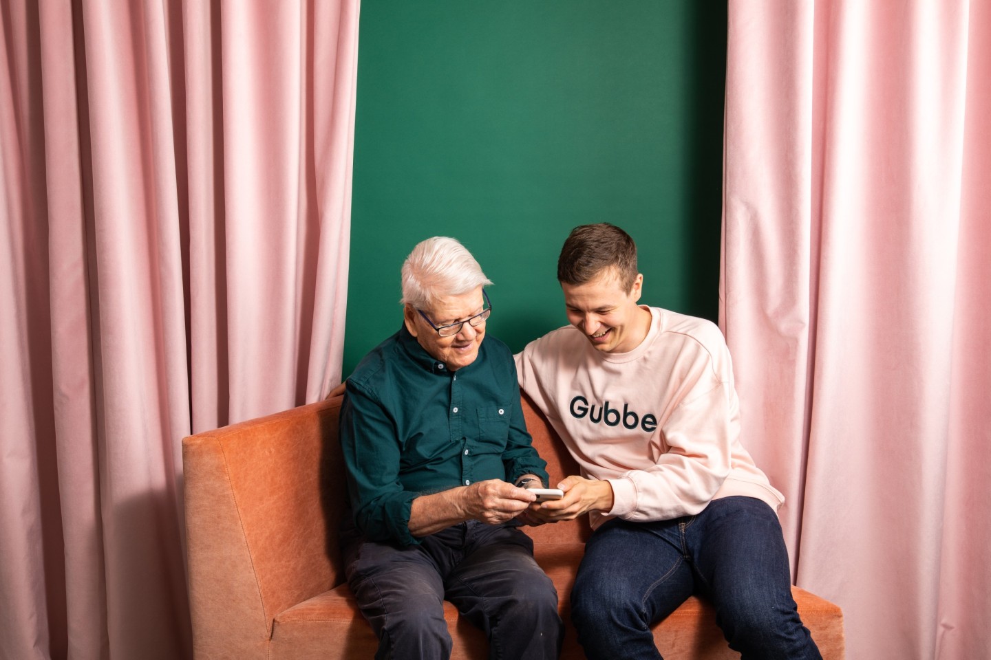 Nuori ja vanha mies istuvat sohvalla ja katsovat yhdessä kännykkää.